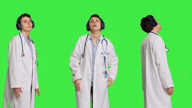 Joyful physician dancing around in the studio with music on headset, having fun with modern cool songs against greenscreen backdrop. Medic woman enjoying audio tunes and singing. Camera B.