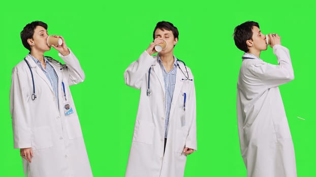 Happy medic serving hot coffee cup as refreshment against greenscreen backdrop, wearing white coat and drinking caffeine beverage. General practitioner enjoying drink, medical expertise. Camera B.
