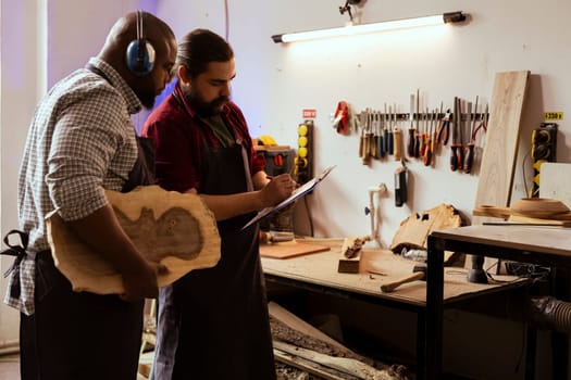 Carpenter and coworker doing verifications on piece of wood before using it for furniture assembly. Teamworking men inspecting lumber block for damages, crosschecking with checklist