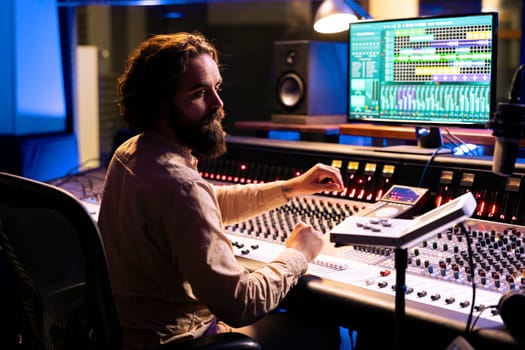 Music technician mixing and mastering songs with motorized faders and knobs, using control desk dashboard and computer. Young sound engineer creating tracks for an album in studio.