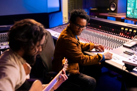 Artist playing a new record on his guitar in control room, recording and producing music with audio engineer in studio. Musician singing on electro acoustic instrument next to mixing console.