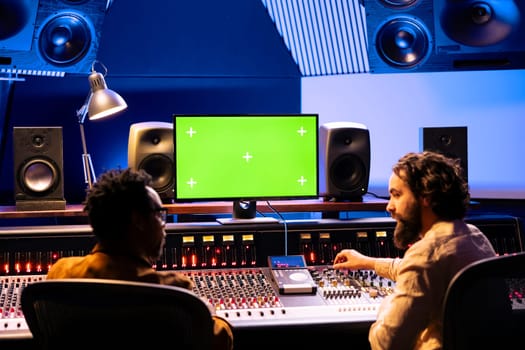 Musicians team working together on mixing tracks with isolated monitor screen, professional recording studio. Artist and audio technician using console and control panel board, knobs and faders.