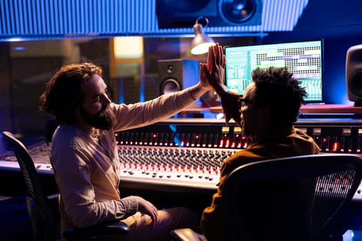 Pleased musician doing high five gesture with producer after finishing musical project, recording and editing music for his new album. Artist and technician enjoy successful records in control room.