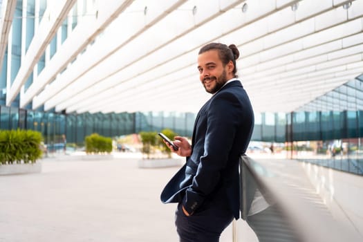 Businessman standing outdoor use smartphone looking camera screen and smile. Handsome hispanic male business person in business suit holding smartphone in hand. Modern business person low angle view