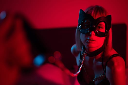 Blonde girl in a leather mask and handcuffs in the bedroom in red blue neon light. Role-playing games
