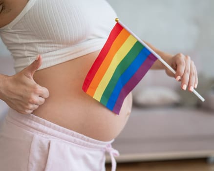 A woman holds a rainbow flag against the background of a naked pregnant belly and shows a thumbs up