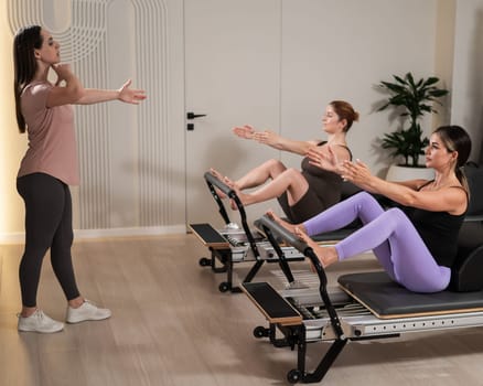 Two pregnant women are doing Pilates on a reformer. The instructor teaches prenatal yoga