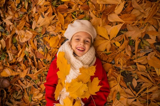 An overhead view of a caucasian girl in a red coat and beret lies on yellow foliage. Walk in the park in autumn