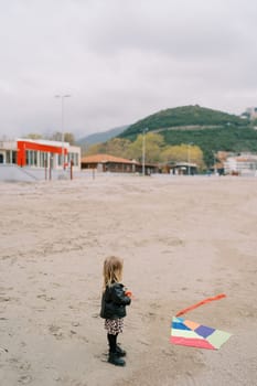 Little girl stands on the beach with a reel from a multi-colored kite on the sand and looks at it. Back view. High quality photo