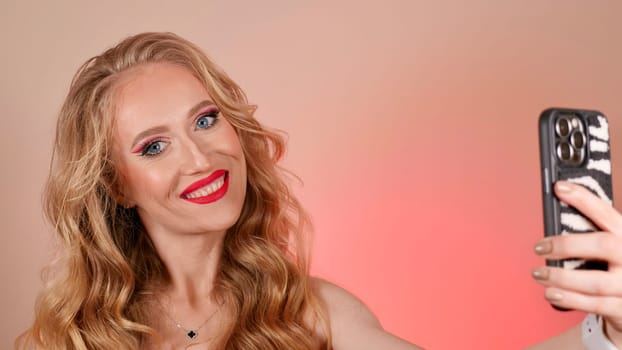 A young woman in a red dress takes a selfie with long blond hair and makeup. The girl is smiling and looking at the camera close-up