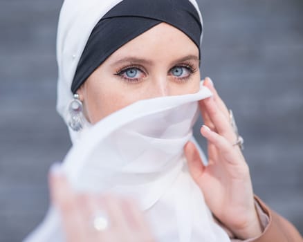 Portrait of a young blue-eyed woman in a hijab against a gray brick wall. Muslim woman with white scarf covering her face
