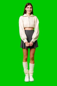 A cheerful young woman stands with confidence against a green backdrop, dressed in a white zippered sweater and a grey skirt. Her hands are gently clasped in front of her as she conveys a friendly demeanor with her smile.