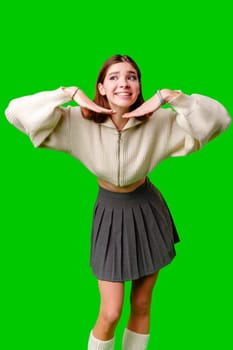 A cheerful young woman is standing against a vibrant green backdrop. Dressed in a cozy white sweater and gray pleated skirt, she playfully pulls at her collar with both hands. Her fashion-forward outfit is complemented by a pair of comfortable white socks, and her bright smile radiates a sense of lively confidence.