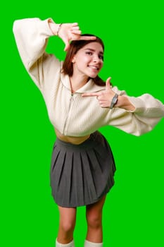A young woman stands against a vibrant green screen, creating a finger frame with her hands as if visualizing a composition or focusing on a specific view. She is dressed in a casual white sweater and a gray pleated skirt, and exudes a playful and creative vibe.