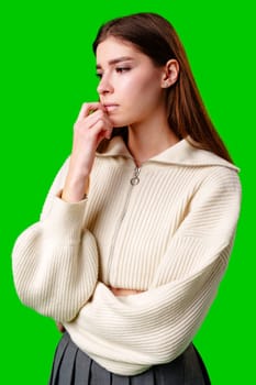 A thoughtful young woman with long brown hair is captured in a moment of contemplation, lightly resting her fingers on her chin. She appears reflective and focused, sporting a casual white zipper sweater, against a stark, vibrant green background that highlights her serene demeanor.