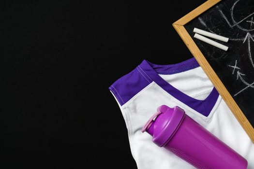A sports uniform with white and purple colors is laid out beside a purple water bottle and a chalkboard with chalk pieces. The scene suggests preparation for a sporting event or a team strategy session, possibly in a locker room or a team meeting area.