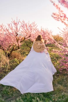 Woman blooming peach orchard. Against the backdrop of a picturesque peach orchard, a woman in a long white dress and hat enjoys a peaceful walk in the park, surrounded by the beauty of nature