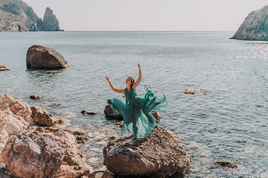 Woman green dress sea. Female dancer in a long mint dress posing on a beach with rocks on sunny day. Girl on the nature on blue sky background