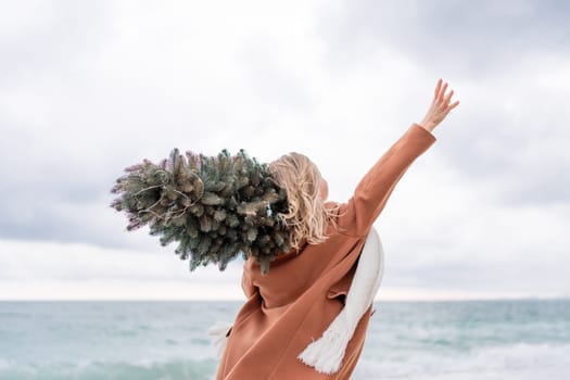 Blond woman Christmas sea. Christmas portrait of a happy woman walking along the beach and holding a Christmas tree on her shoulder. She is wearing a brown coat and a white suit