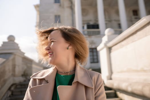 A woman with long hair is standing on a set of stairs. She is wearing a green shirt and a tan coat