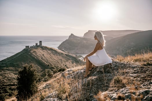 A woman is sitting on a hillside overlooking the ocean. She is wearing a white dress and has blonde hair. The scene is serene and peaceful, with the ocean in the background