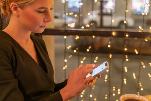 A woman is looking at her cell phone while sitting in a window. The scene is set in a city at night, with lights and a car visible in the background