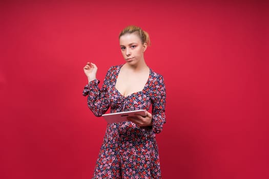 A woman in a purple floral dress is holding a tablet computer, her arm raised in a gesture. She has a microphone pinned to her onepiece garment, with lipstick and flash photography visible