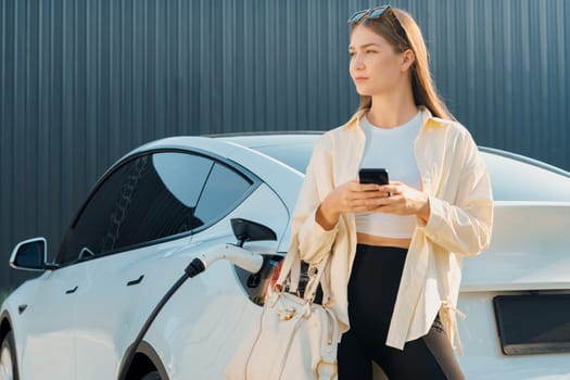 Young woman holding shopping bag and use smartphone to pay for electricity for recharging EV car battery from charging station at city mall parking lot. Modern woman go shopping by eco car. Expedient