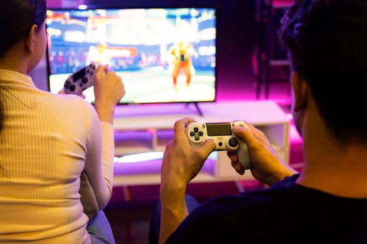 Couple joyful player video game on TV using joysticks at back side, playing fighting game with winner together raising fist up in studio room in red neon light at comfy living home place. Postulate.