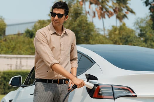 Young man recharge electric car's battery from charging station in outdoor green city park in springtime. Rechargeable EV car for sustainable environmental friendly urban travel lifestyle. Expedient