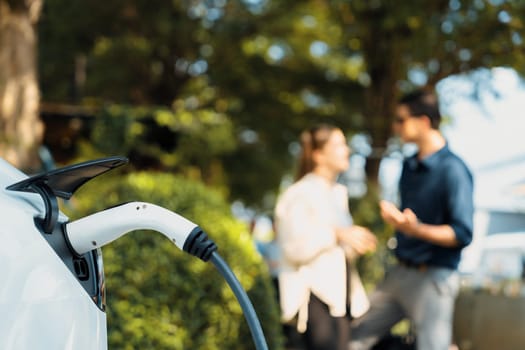 Young couple recharge electric car's battery from charging station in outdoor green city park in springtime. Rechargeable EV car for sustainable environmental friendly urban travel lifestyle.Expedient