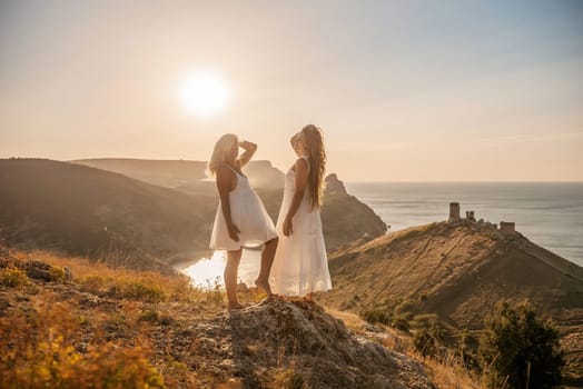 Two young girls are standing on a hillside, one of them wearing a white dress. The sun is shining brightly, creating a warm and inviting atmosphere. The girls seem to be enjoying their time together