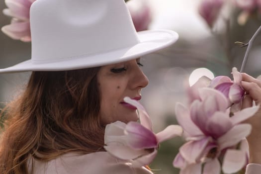 Woman magnolia flowers, surrounded by blossoming trees, hair down, white hat, wearing a light coat. Captured during spring, showcasing natural beauty and seasonal change