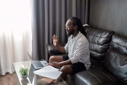 African American man working with laptop computer video calling and conference, remote work while sitting at sofa in living room. Black guy do freelance work at home office.