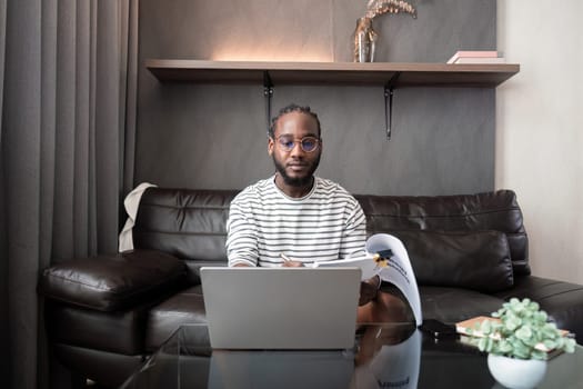 African American man working with laptop computer remote while sitting at sofa in living room. Black guy do freelance work at home office.