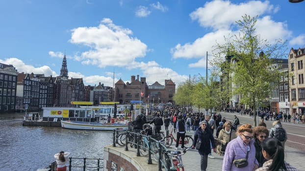 Amsterdam Netherlands 21 April 2024, A diverse group of people stroll down a charming street beside a peaceful river, taking in the scenery and chatting animatedly.