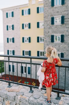 Little girl stands leaning on a fence and looks at an apartment building with a playground. Back view. High quality photo