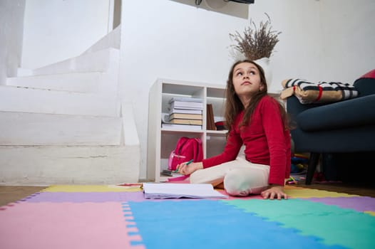 Adorable Caucasian little child girl in home clothes, sitting on a colorful puzzle carpet, dreamily looking aside, solving mathematics problems, writing essay while doing homework in her children room