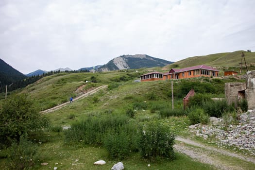 A small wooden home with a red roof on a hill, overlooking a valley and mountains. Blue sky, white clouds, green grass.