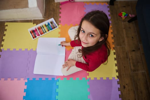 View from above of a cute little child girl smiling looking at camera, drawing with pastel pencils, sitting on a colorful puzzle carpet in cozy domestic room. People. Childhood. Art and creativity