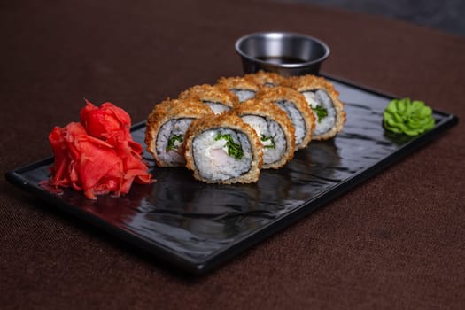 Deep-fried sushi roll with chopsticks and soy sauce on a black plate, made with rice, seaweed, fillings, and golden brown.