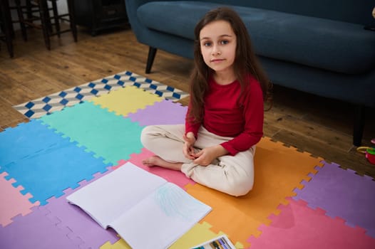 Adorable elementary age school child, little girl at home, doing homework, writing on a copy book, smiling looking at camera, sitting on a puzzle carpet in a cozy home interior. Education concept