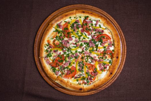 Delicious pizza with ham, tomatoes, bell peppers, and fresh herbs on a wooden board. Isolated on a brown background.