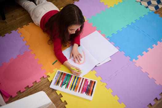 Overhead view of a talented little child, school girl taking out colorful pencil from pencil case, drawing beautiful cloud with rainbow, lying on a multi colored puzzle carpet in cozy home interior.