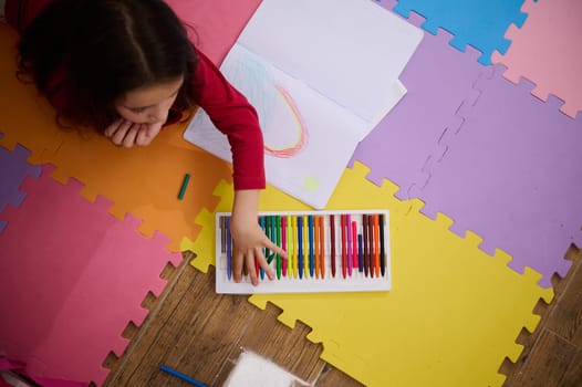 View from above of a little child girl taking out colorful pencil from pencil case, drawing beautiful cloud with rainbow, lying on a multi colored puzzle carpet in cozy home interior. Kids education