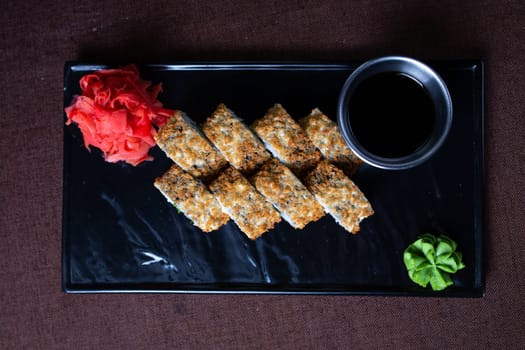 Deep-fried sushi roll with chopsticks and soy sauce on a black plate, made with rice, seaweed, fillings, and golden brown.