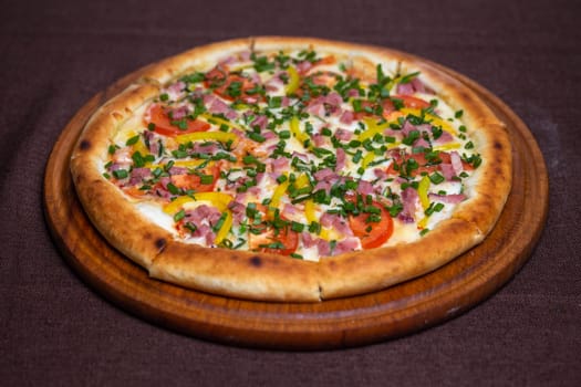 Delicious pizza with ham, tomatoes, bell peppers, and fresh herbs on a wooden board. Isolated on a brown background.