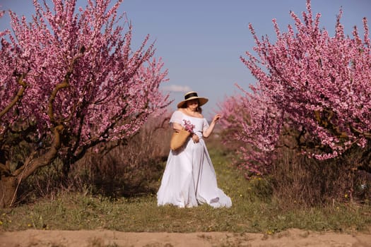 Woman blooming peach orchard. Against the backdrop of a picturesque peach orchard, a woman in a long white dress and hat enjoys a peaceful walk in the park, surrounded by the beauty of nature