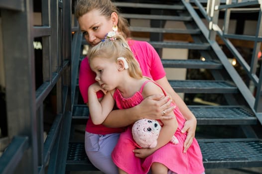 Blonde little girl with cochlear implant playing with her mother outdoor. Hear impairment deaf and health concept. Diversity and inclusion. Copy space.