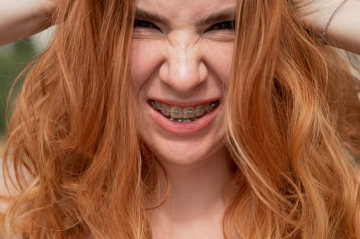 Close-up portrait of a young red-haired woman with braces on her teeth. Girl makes faces at the camera outdoors